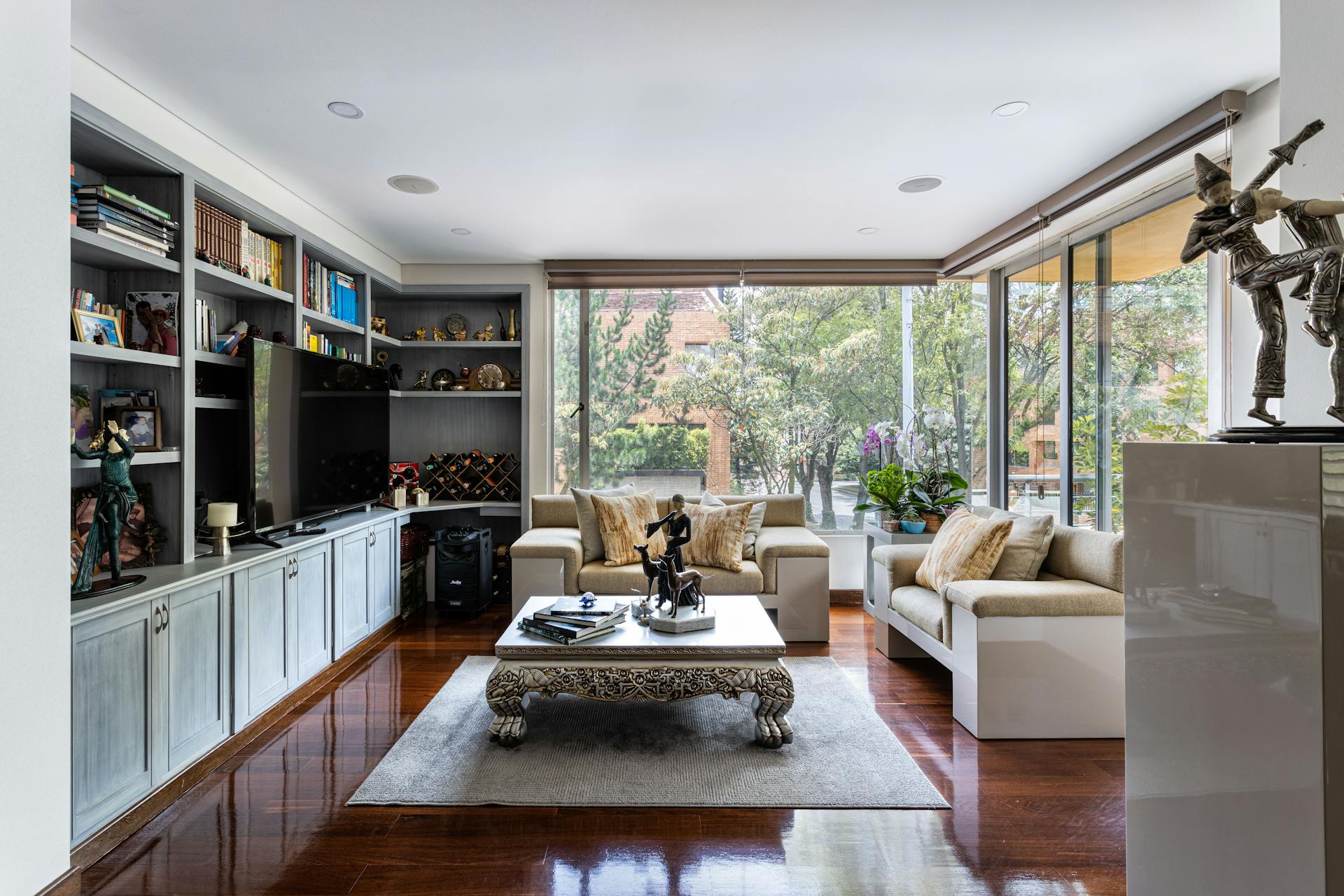 Sun-filled living room with large windows, motorized shades raised to let in full afternoon light, tree views, warm wood floors, and a built-in shelving wall
