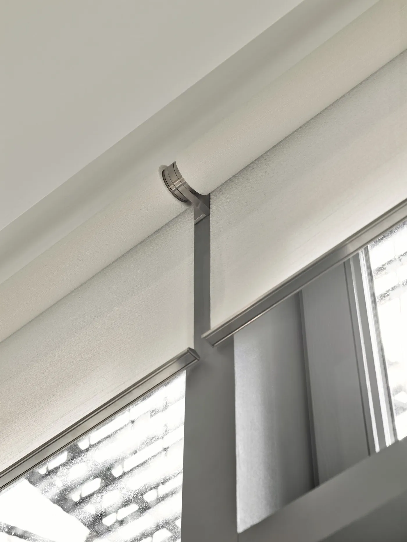 Close-up looking up at two motorized roller shades mounted side by side on a window frame, sharing a brushed nickel center bracket — clean outside mount installation
