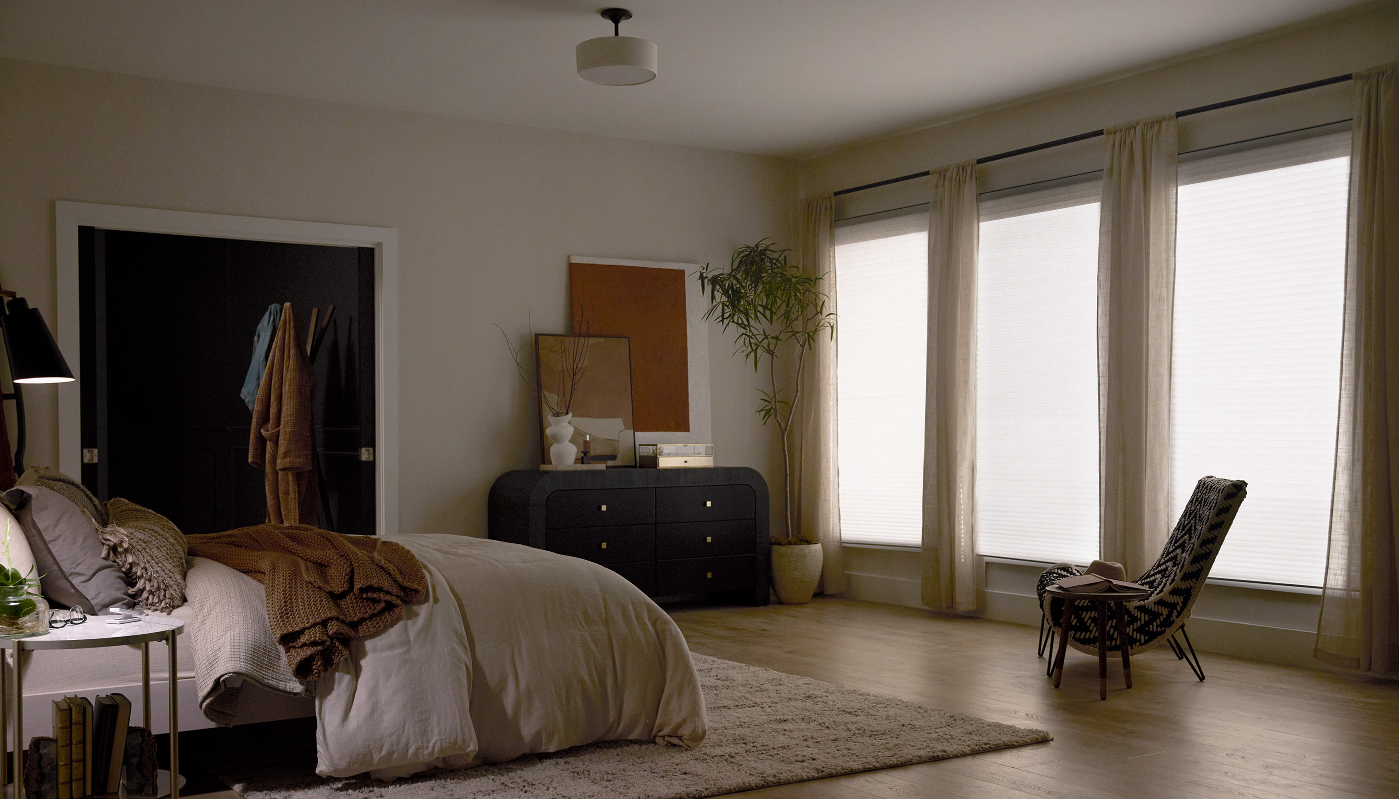 Moody primary bedroom at dusk with motorized roller shades fully lowered on three tall windows, sheer curtains, warm bedside lamp — the room set perfectly for evening