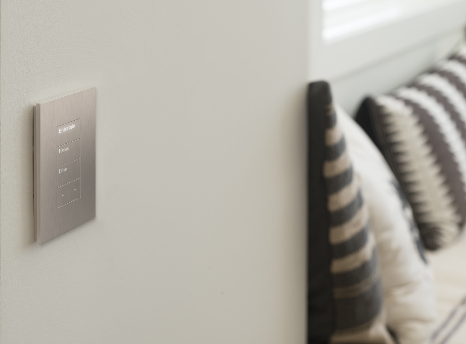 Lutron scene keypad on a neutral wall with brushed aluminum finish, showing Entertain, Relax, and Dine buttons with raise and lower controls — soft pillows visible in the background